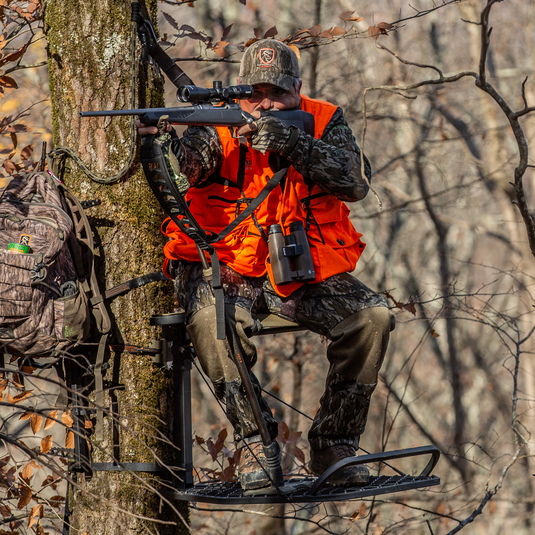 A hunter uses a Primos Gen3 Tall Monopod Trigger Stick to aim a rifle from a tree stand in the woods.