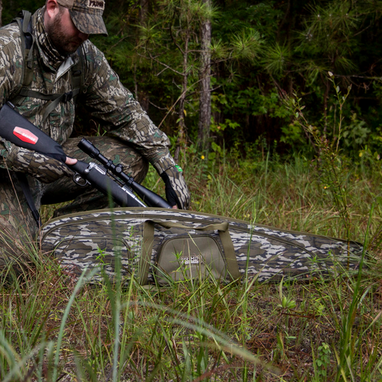 A person kneels outdoors, holding a rifle beside a Primos Mossy Oak Bottomland Scoped Rifle Case on the grass.