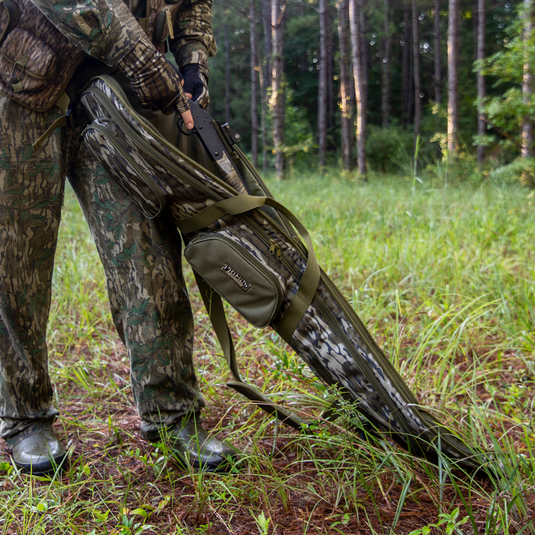 Lower body in Mossy Oak Bottomland holds Primos Shotgun Case in a grassy forest clearing.