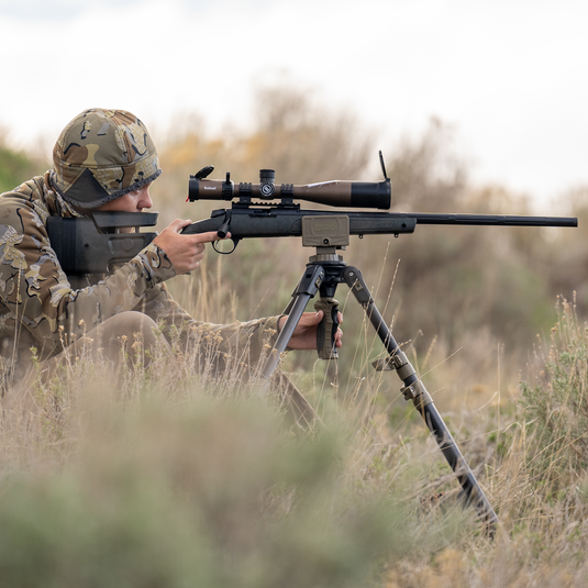 A person in camouflage aims a rifle with a scope on the Primos Trigger Stick Apex Carbon Fiber Tripod outdoors.