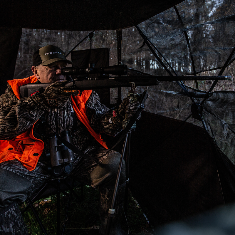 Load image into Gallery viewer, A hunter in camo and orange vest aims a rifle with the Primos Gen3 Tall Bipod Trigger Stick in a woodland blind.
