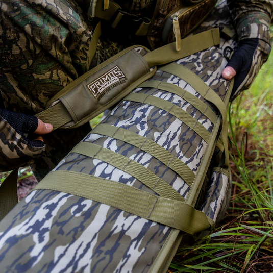 A person holds a Primos Mossy Oak Bottomland Shotgun Case with choke tube holders in a grassy outdoor area.