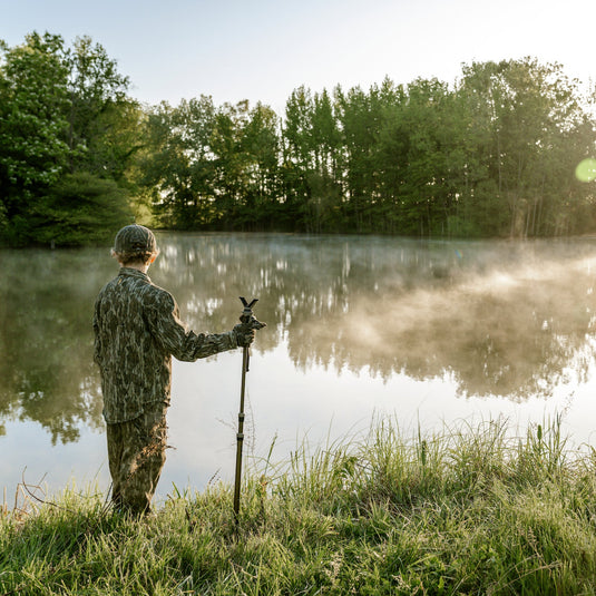 Person in camouflage standing by a lake with trees in the background