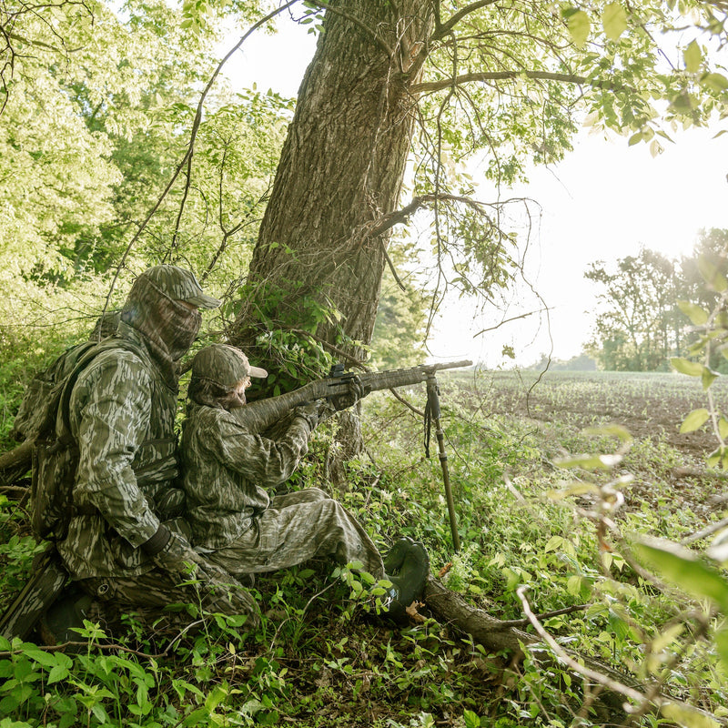 Load image into Gallery viewer, Two hunters in camouflage sitting in a forest with one holding a rifle.
