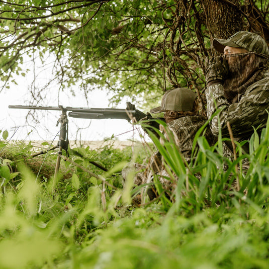 Two people in camouflage gear in a forest setting with one holding a rifle.