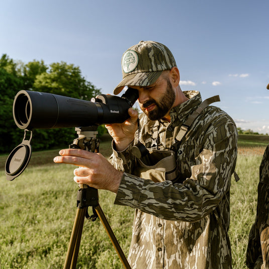 Two people in camouflage clothing using a telescope in an open field.