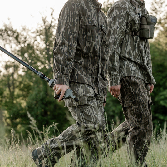 Two hunters in camouflage walking through a grassy field with trees in the background.