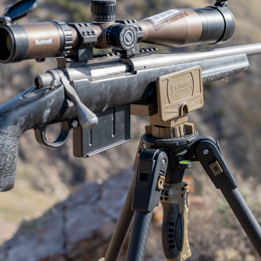 A Primos Trigger Stick Apex Carbon Fiber Tripod holds a rifle with a scope outdoors in rocky, mountainous terrain.