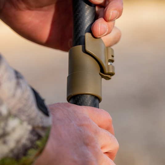 A person adjusts a brown clamp on a Primos Gen3 Short Bipod Trigger Stick outdoors.
