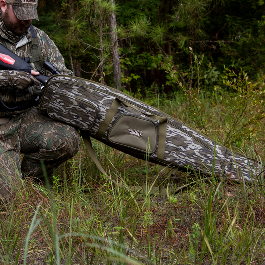 A man holds a Primos Mossy Oak Bottomland Scoped Rifle Case while standing in the woods.