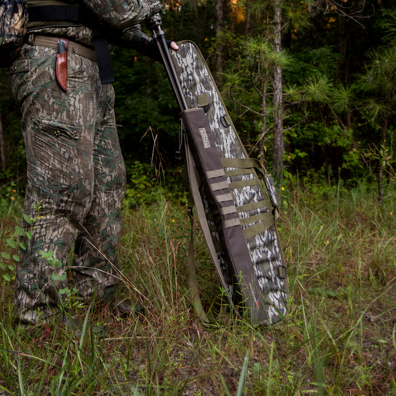 Load image into Gallery viewer, A person in camo stands in grass holding a Primos Mossy Oak Bottomland Scoped Rifle Case in a wooded area.
