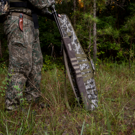 A person in camo stands in grass holding a Primos Mossy Oak Bottomland Scoped Rifle Case in a wooded area.