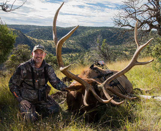 Smiling hunter kneels beside a large elk with antlers in grassy, hilly landscape under a partly cloudy sky.