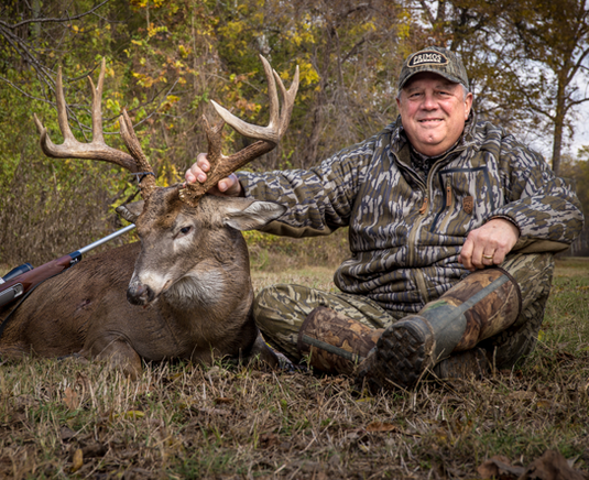 A smiling hunter in camouflage sits on grass, holding the antlers of a large buck deer he has hunted.