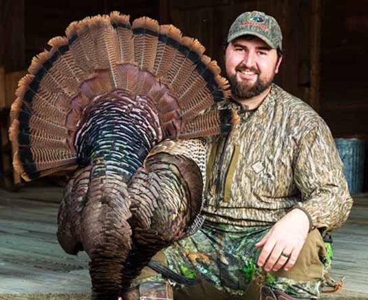 A smiling man in camouflage poses with a large wild turkey displaying its feathers outdoors.