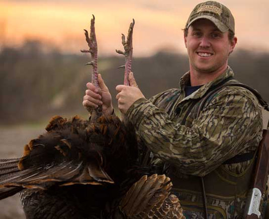 Smiling hunter in camouflage holding up a wild turkey by its legs outdoors at sunset.