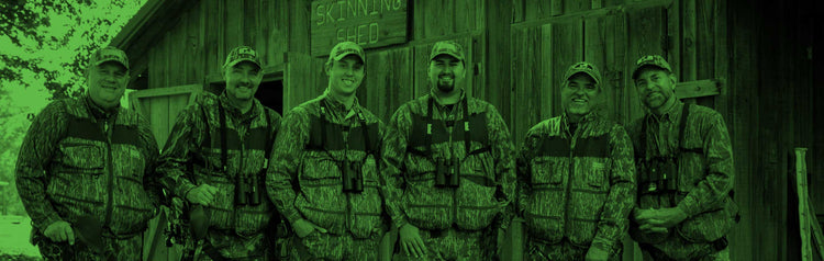 Six men in camouflage gear stand smiling in front of a wooden building, filtered by green night vision.
