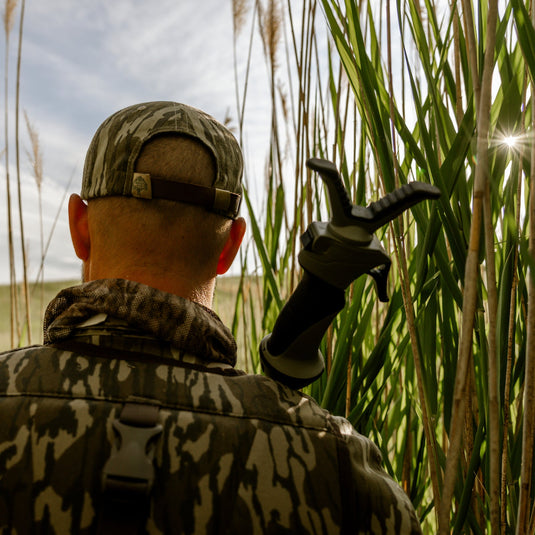 Person in camouflage clothing holding a shotgun, standing among tall grasses.