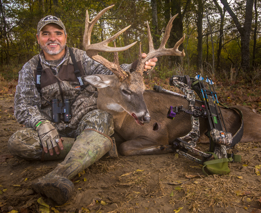 Smiling hunter in camouflage sits on ground, holding antlers of a large deer with a bow and arrows beside him in the woods.