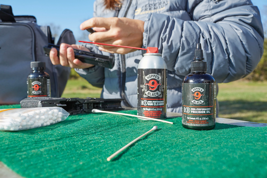 Person preparing a gun cleaning kit with various Hoppe's Black gun cleaner and oil bottles and swabs on a green mat outdoors.