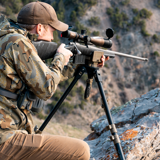 A person in camouflage aims a rifle with a scope on a Primos Trigger Stick Apex Carbon Fiber Tripod near rocks.