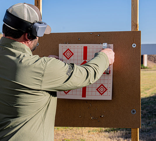 A person attaches a paper target to a board outdoors, wearing ear protection and a cap.