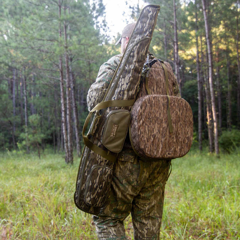 Load image into Gallery viewer, A hunter with a Primos Mossy Oak Bottomland Shotgun Case walks through a tall forest carrying a backpack.
