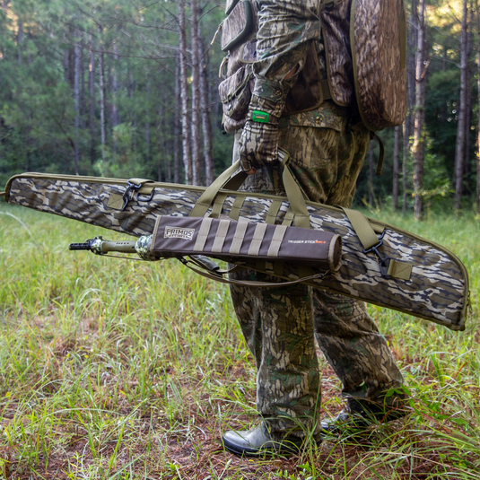 Person in Mossy Oak Bottomland holds a Primos shotgun case with choke tube holders and a hunting backpack in the forest.