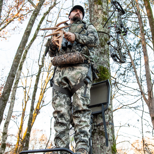 A hunter in camouflage holds Primos Fightin' Horns deer rattling antlers on a tree stand, compound bow nearby.