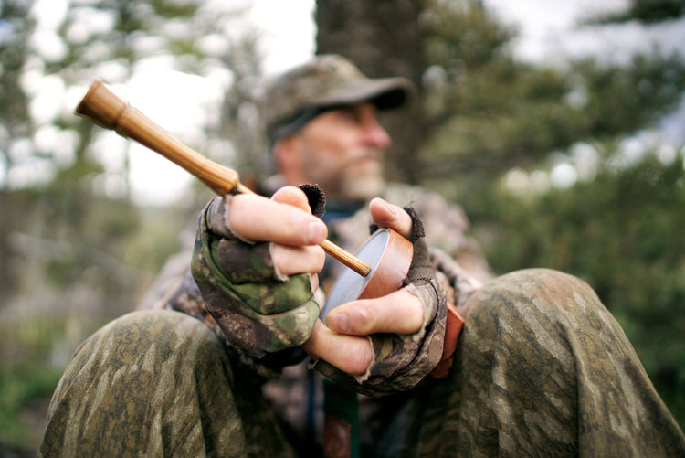A hunter in camouflage holds a turkey call, sitting outdoors among trees, with a blurred background.