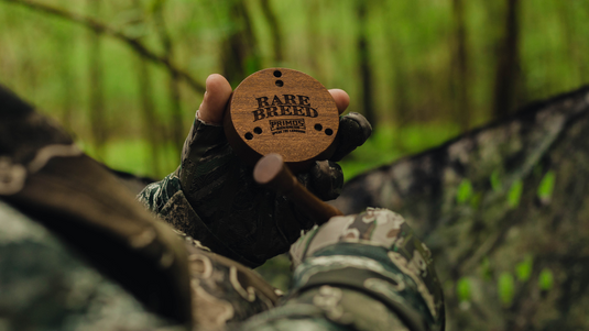 A gloved hand holds a Rare Breed Primos turkey call in a green, wooded outdoor setting.