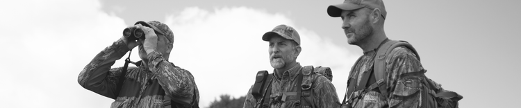Three men in camouflage gear outdoors; one uses binoculars while the others look ahead, cloudy sky in background.