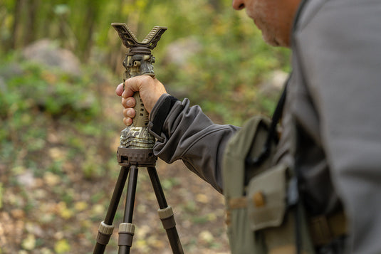 Person outdoors adjusting a camouflage-patterned tripod with a V-shaped mount, surrounded by greenery.